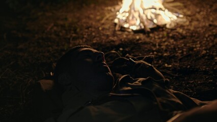 Soldier sleeping on the ground near a blazing campfire at night in a close up view as he turns...