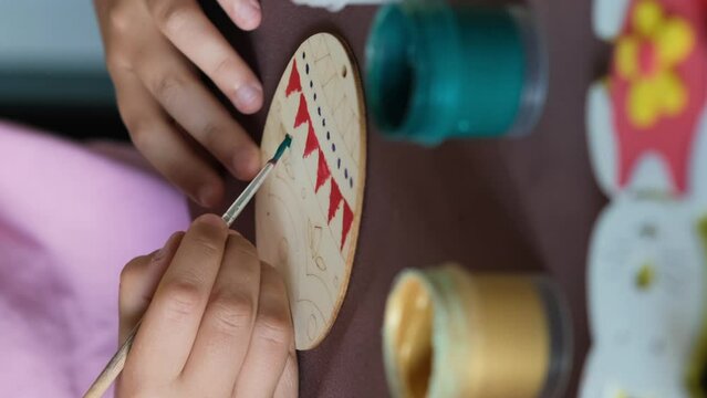Vertical video. Close-up of hand of child girl painting wooden egg fro Easter at table