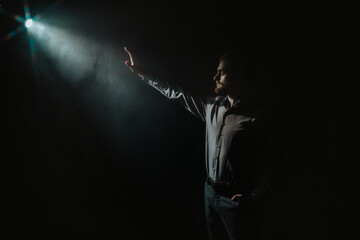 A young man with a beard extends his hand to a ray of light on a black background.