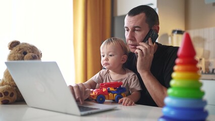 father working from home a remotely with baby son in his arms. pandemic remote work business concept. father tries to work at home in kitchen, baby children interfere sitting on their fun hands
