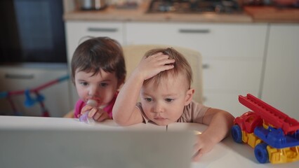 twin baby children a playing laptop watching video in the kitchen. happy family kid dream concept. baby lifestyle twins playing video game on laptop looking at screen in kitchen