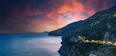 Amalfi Coast, Italy. View over Praiano on the Amalfi Coast at sunset. Street and house lights at dusk. In the distance the island of Capri on the horizon. Amalfi Coast road. Banner header image.. - obrazy, fototapety, plakaty