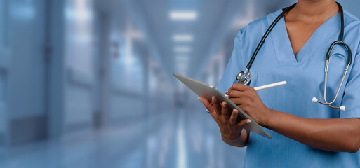 A focused nurse in blue scrubs using a digital tablet while standing in a brightly lit hospital