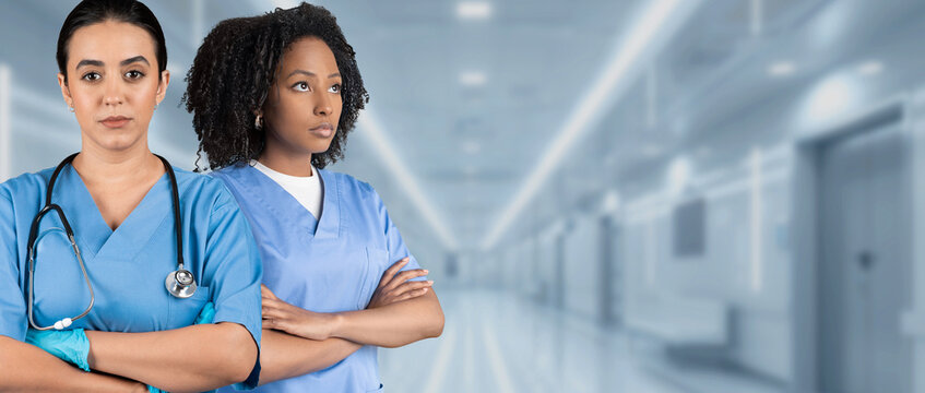 Two serious female nurses in blue scrubs with crossed arms and stethoscopes