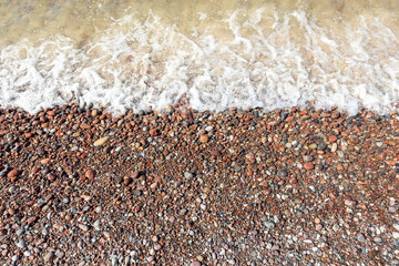 A white foaming sea wave washes the brown-red pebbles of the coast.