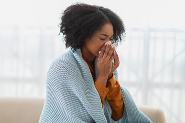Sick woman with curly hair sneezing into a tissue, wrapped in a comforting blue blanket