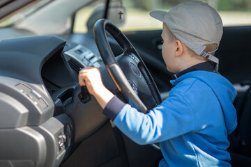 Cute little boy behind the wheel of an fathers car, he holds the steering wheel, happily looks to...