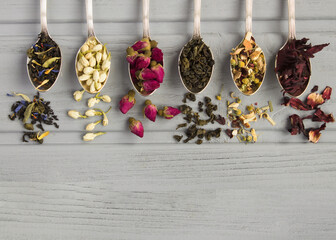 Different dried tea in the silver spoons on the gray wooden  background. Top view. Copy space.