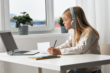 Smiling girl learning at home with a laptop elearning in action The cute, young Caucasian schoolgirl sits at a desk, immersed in her online education With headphones on, she engages in a virtual