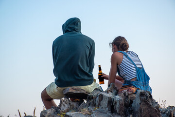 Boy in a sweatshirt and girl with blue suspenders having a beer sitting on a rock