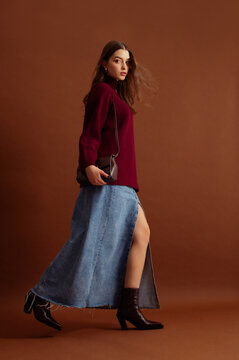 Fashionable confident woman wearing burgundy color knitted sweater, blue denim maxi skirt, leather pointed toe ankle cowboy boots, posing on brown background. Full-length studio fashion portrait