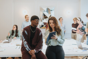 Young coworkers, a man and woman, deep in discussion holding a tablet with co-workers interacting...