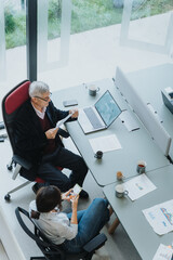 Elevated view of a senior businessman analyzing paperwork while his young female colleague uses a smart phone. They are working at a modern office workspace with technology and daylight.