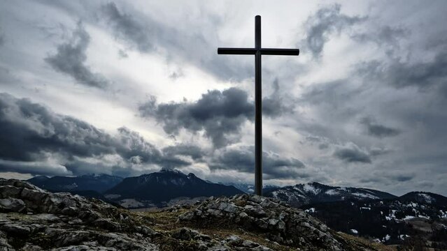 Christian cross on a rock in a mountain landscape, Dramatic clouds over the silhouette of the mountains in winter, Timelapse