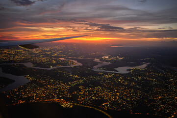 Vista da janela de avião comercial do Rio Iguaçu e Rio Paraná durante voo noturno com luzes da cidade e céu dramático em tons vermelho, roxo e azul. Foz do Iguaçu, Paraná. 2024.