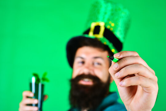 Patrick's day celebration. Bearded man in leprechaun hat and glass green beer with green marker. Selective focus. Man's hand with green felt-tip. School supplies. Painting. Copy space for advertising.