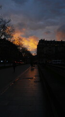 Grande avenue ou allée urbaine en début de soirée, couché de soleil derrière ou à travers de haut bâtiment parisien, façade gothique, ciel bleu foncé et nuages jaunes, beauté photographique lumière,