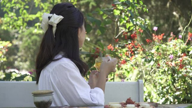 A beautiful asian woman in white is sipping a glass of hot tea in the morning with bokeh garden backgrounds. Herbal drinks, Alternative medicine, Healthy food, Relaxation, Leisure activity concept