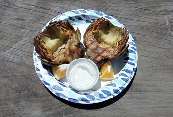 Grilled globe artichokes with dipping sauce and orange wedges on paper plate