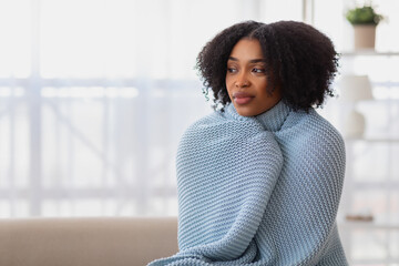 Pensive African American woman with curly hair wrapped snugly in a blue knitted sweater - obrazy, fototapety, plakaty