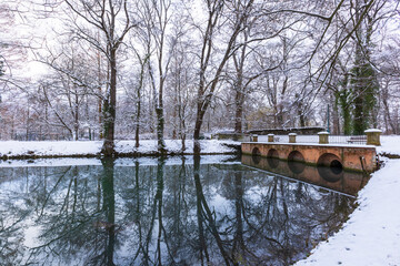 Winter snowy landscape. A brick bridge leading over a calm river, which is lined with tall snow-covered trees reflecting on the surface of the river.