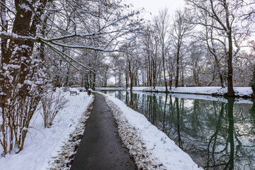 Winter snowy landscape. A small road leads in the middle, to the left is a river with a calm surface in which snow-covered trees are reflected.