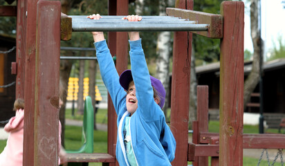 Fototapeta na wymiar A young girl dressed in a blue hoodie and purple hat reaches up as she plays on a wooden jungle gym structure in a verdant park setting