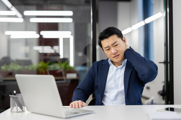 Fototapeta premium Overworked overtired asian man in business suit at workplace inside office, businessman has severe pain in neck, massaging muscle , long time sitting work with laptop, daily business of financier.