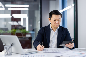 Serious thinking businessman accountant checking financial reports, mature concentrated asian man behind paper work, calculator in hands, worker in business suit with laptop inside office.