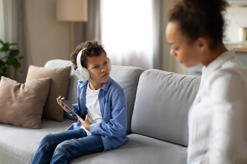 Conflicted young boy with headphones and tablet, mother looking concerned