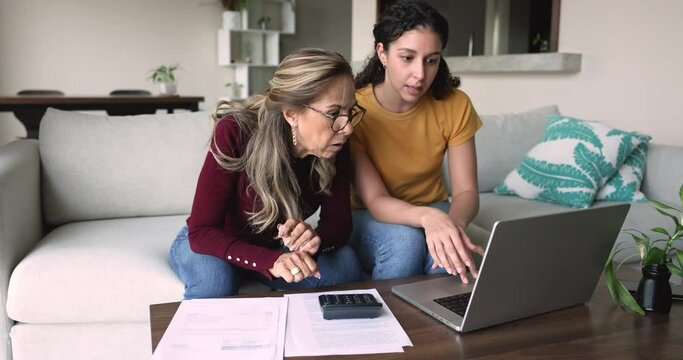 Grown up daughter helps with family budget management to mature mother, family sit on sofa pay bills using on-line bank system on laptop, counting savings and outcomes, analyze expenses, make payments