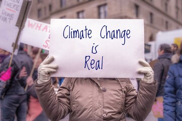 Hands of protester holding homemade sign reading "Climate Change is Real" during demonstration about global warming in urban area. Focus on foreground with intentional background blur.
