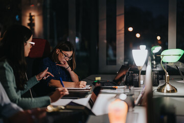 Two focused female colleagues collaborate at a late-night business meeting in a well-lit office setting, indicating teamwork and dedication.