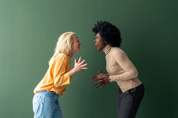 Two young interracial women yelling at each other against a green background in a studio.