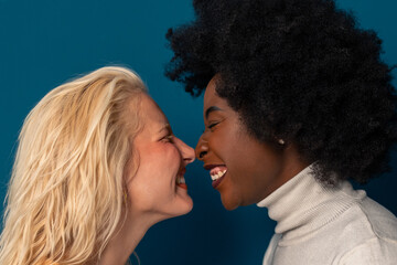 Close up of a cheerful young women with different skin tones standing together face to face in a studio. Headshot of a two diverse women with noses touching against a blue background.