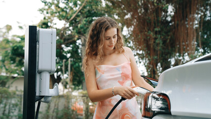 Closeup woman recharge EV electric car's battery at parking lot in natural green park. Clean energy...