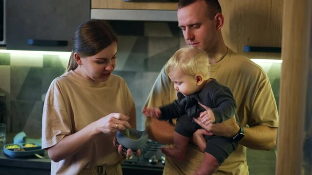 Man is holding adorable baby boy with blond hair. Woman is mixing food in the bowl to cool it and hungry kid wants to reach the dish.
