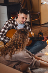 Obraz premium Young handsome man musician playing guitar instrumental music for his happy beautiful girlfriend near fireplace in a countryside cottage house, mountain chalet. Winter or autumn, cozy home atmosphere
