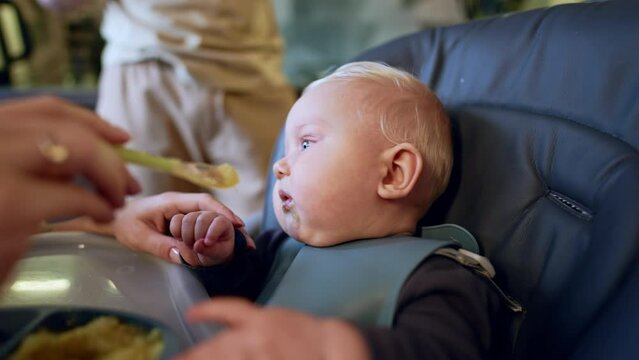 Chubby little infant starting to cry at the high chair. Mom gives a spoon with food to her child. Close up.