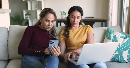 Young adult daughter sit on sofa at home with laptop helping to elderly mother with data backup, synchronization, new application or gadget usage. Multigenerational family using modern tech, lifestyle