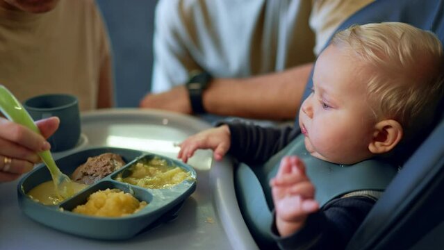 Infant baby boy with plump cheeks and blond hair sits at high chair. Mom feeds the child from spoon. Kid sneezes during meals.