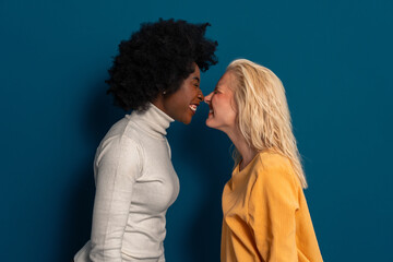 Two cheerful young women with different skin tones standing face to face with noses touching in a...
