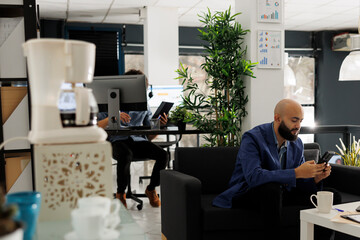 Arab business worker chatting with coworker using smartphone while working in modern open space. Marketing professional sitting on couch and texting in social media on mobile phone in start up office