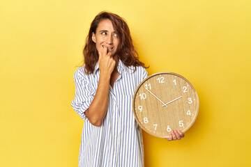 Middle aged woman holding a wall clock on a yellow backdrop relaxed thinking about something...