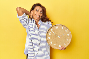 Middle aged woman holding a wall clock on a yellow backdrop touching back of head, thinking and...