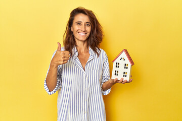 Middle aged woman holding a miniature house on yellow backdrop smiling and raising thumb up
