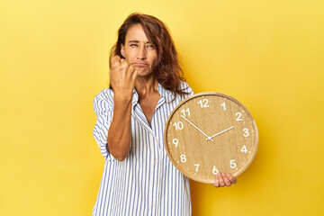 Middle aged woman holding a wall clock on a yellow backdrop showing fist to camera, aggressive...