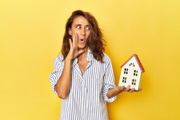 Middle aged woman holding a miniature house on yellow backdrop is saying a secret hot braking news...