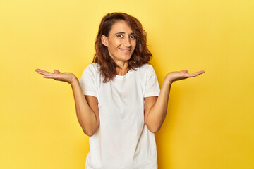Middle-aged woman on a yellow backdrop makes scale with arms, feels happy and confident. - obrazy, fototapety, plakaty