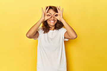Middle-aged woman on a yellow backdrop showing okay sign over eyes